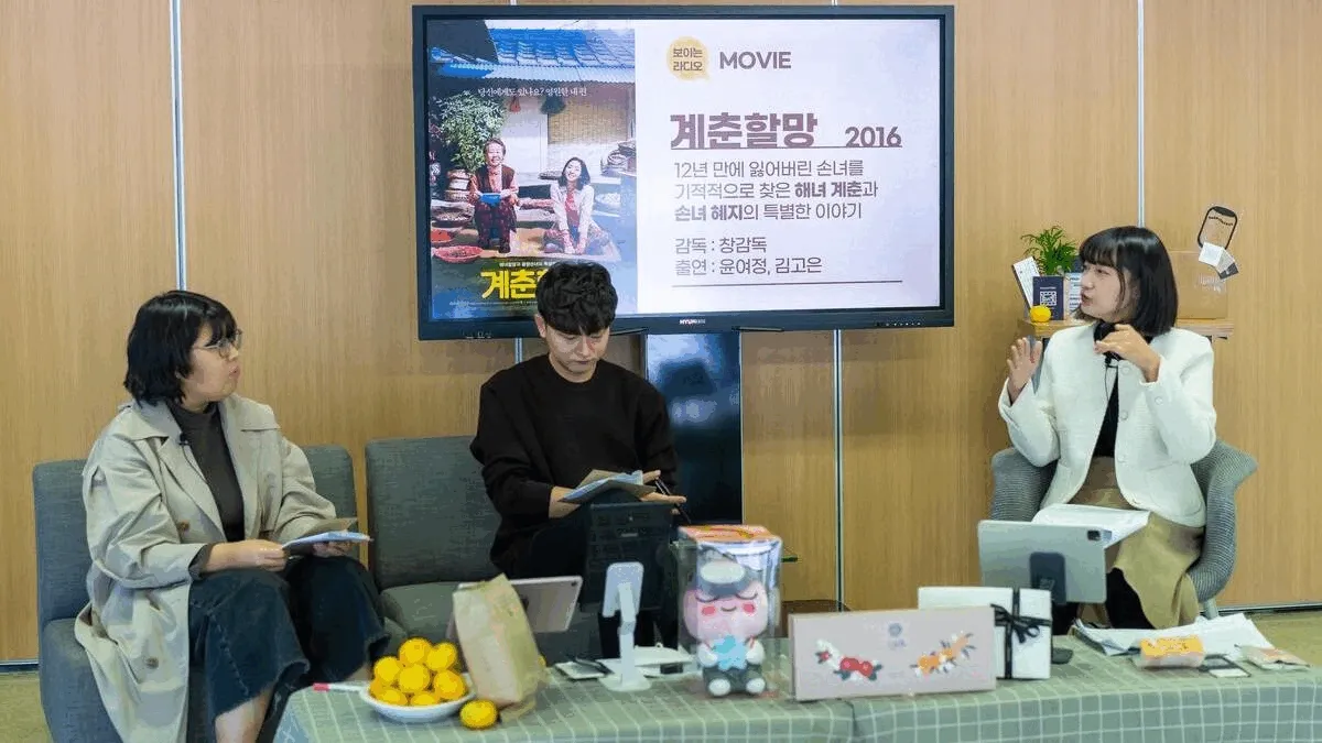 보이는 라디오 진행 — 화면 "보이는 라디오 MOVIE 계춘할망 2016 / 12년 만에 잃어버린 손녀를 기적적으로 찾은 해녀 계춘과 손녀 혜지의 특별한 이야기 / 감독 : 창감독 / 출연 : 윤여정, 김고은", 진행자 3명과 테이블 위 귤·선물