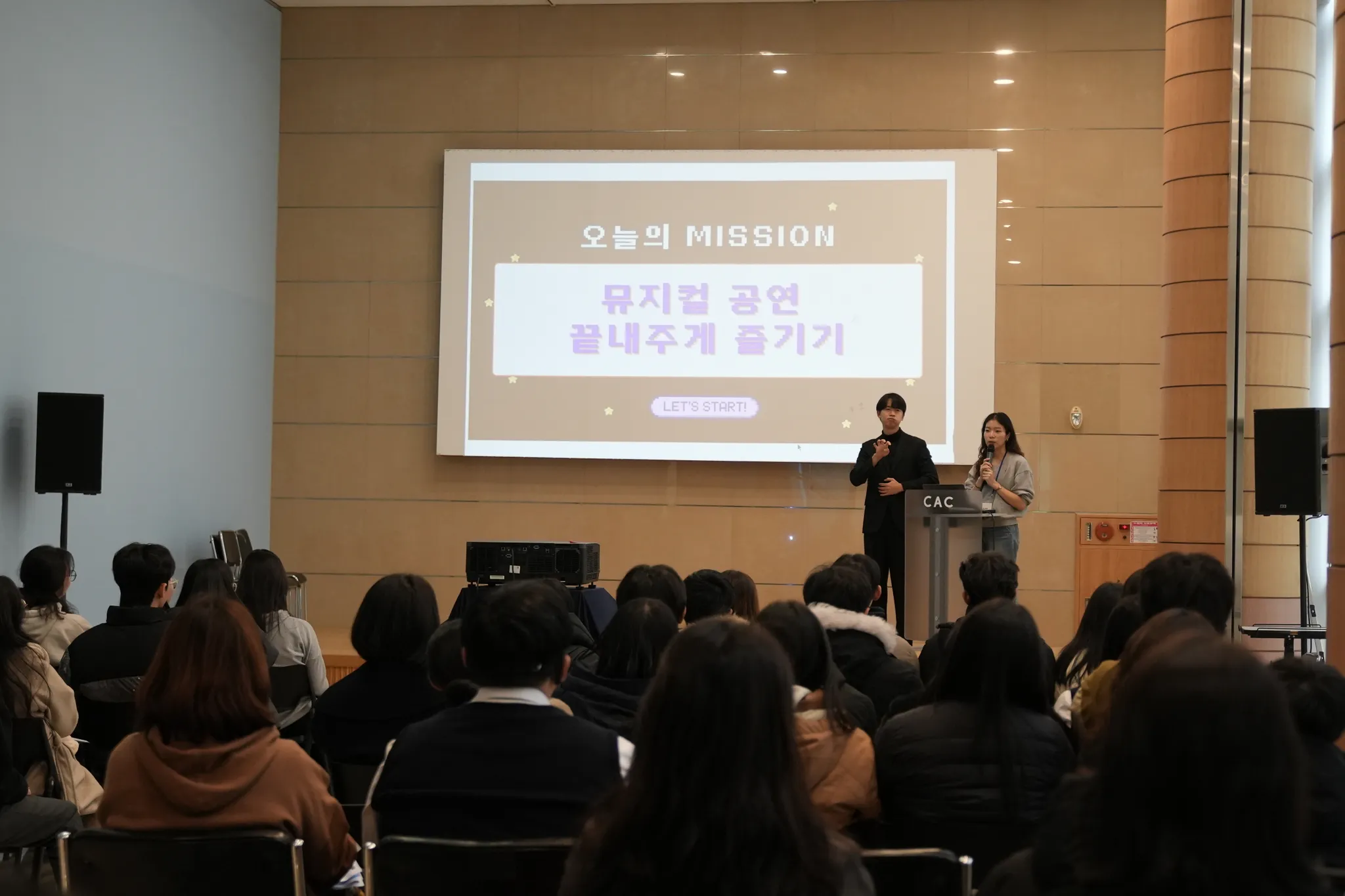 사전 안내 시간 — 화면에 "오늘의 MISSION" 슬라이드, 진행자(마이크)와 수어통역사, 객석의 학생들 뒷모습