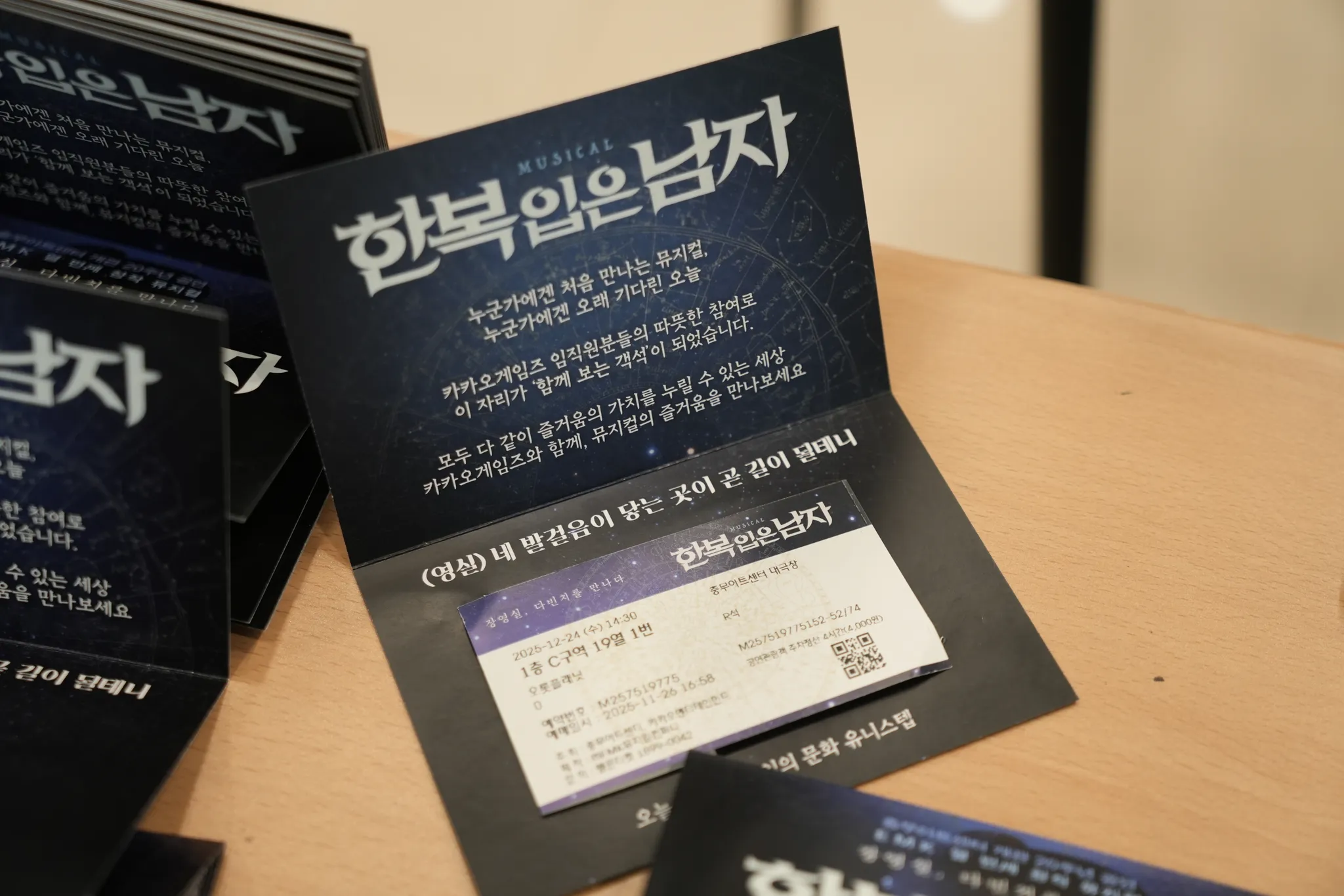 테이블 위 <한복 입은 남자> 안내 카드와 티켓 — "누군가에겐 처음 만나는 뮤지컬, 누군가에겐 오래 기다린 오늘 / 카카오게임즈 임직원분들의 따뜻한 참여로 이 자리가 '함께 보는 객석'이 되었습니다. / 모두 다 같이 즐거움의 가치를 누릴 수 있는