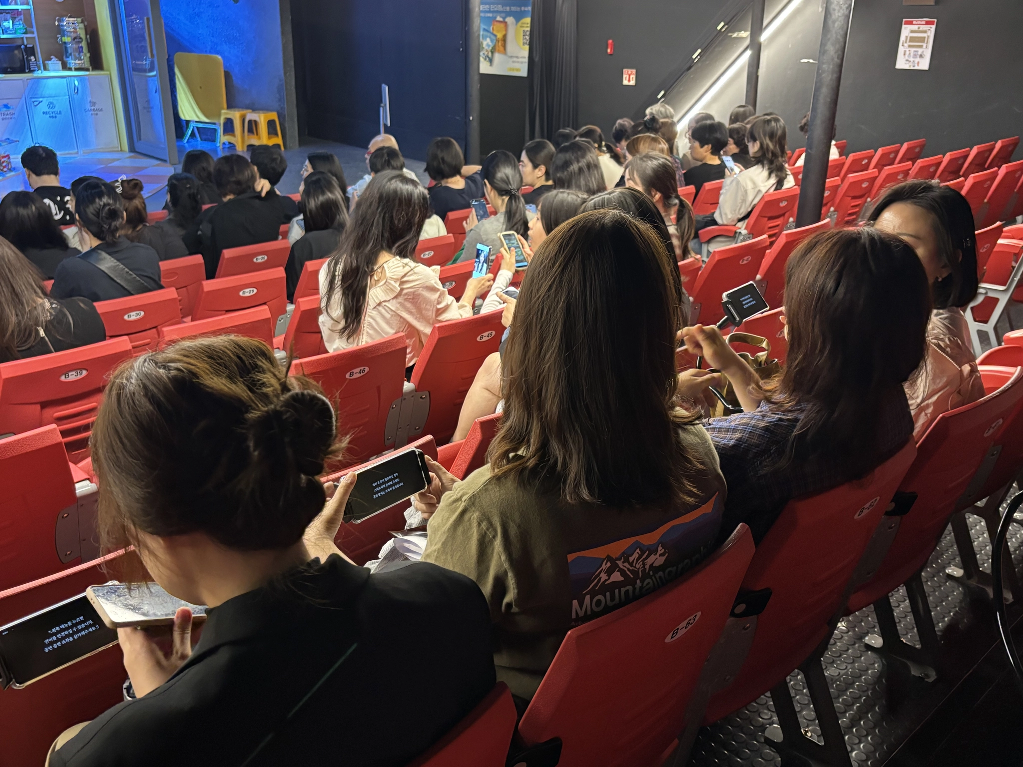 소극장 객석에서 휴대폰·거치대 자막 단말기를 보는 관객들 뒷모습(화면 안내 문구는 작아서 [판독 불가])