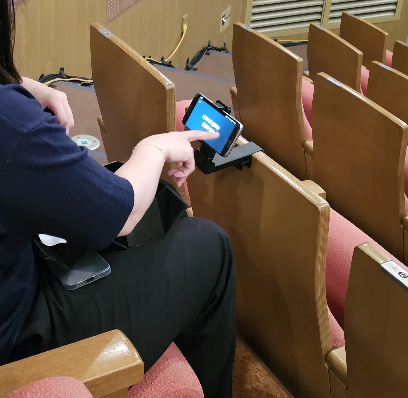 공연장 좌석 등받이에 설치한 거치대 휴대폰 화면을 손가락으로 가리키는 사람 (화면 글씨 [판독 불가])